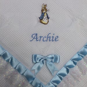 May include: White baby blanket featuring a Peter Rabbit embroidery and the name "Archie" in blue. It has a blue satin ribbon trim and bow. The blanket has a quilted pattern. The blanket is suitable for a baby.