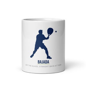 May include: A white ceramic mug featuring a dark blue silhouette of a padel player hitting a ball with a racket. Below the figure, the text reads "BAJADA" in bold blue letters, followed by the smaller text "OFF THE GLASS, STRAIGHT BACK AT THEM".