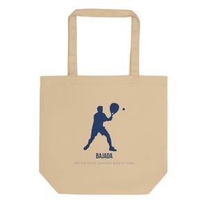 May include: A beige tote bag featuring a dark blue silhouette of a person playing padel with a racket and ball. Below the figure, the text reads BAJADA and OFF THE GLASS, STRAIGHT BACK AT THEM in dark blue and grey capital letters respectively.