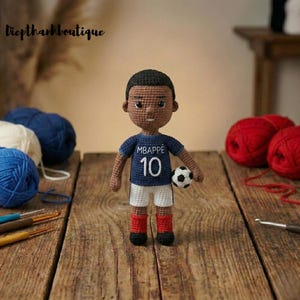 May include: Crocheted doll of a soccer player wearing a blue jersey with "MBAPPE" and the number 10. The doll has white shorts, red socks, and black shoes, holding a soccer ball. The doll is on a wooden surface.
