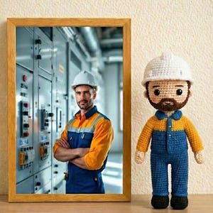 Könnte beinhalten: Ein gerahmtes Foto eines Mannes mit Helm und Arbeitsuniform steht neben einer gehäkelten Puppe, die ein ähnliches Outfit trägt. Die Puppe trägt einen weißen Helm, blaue Latzhosen und ein gelbes Hemd. Das gerahmte Foto hat einen Holzrahmen.