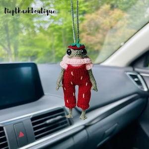 May include: A handmade crochet frog car charm. The frog is wearing a red strawberry-themed outfit with white seed accents, a pink collar, and a green strawberry hat. The charm hangs from a green string. The text "Diepthanhboutique" is in the top left corner.
