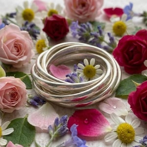 May include: Silver bangles, nested together, are the focal point, set amidst a floral arrangement. Pink and red roses, daisies, and lavender are scattered around the bangles. The scene is a vibrant display of nature and jewellery.