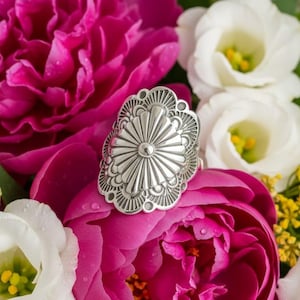 May include: A silver-colored ring with a detailed, sunburst-like design. The ring is set against a backdrop of vibrant pink and white flowers, creating a striking contrast. The ring's intricate patterns and polished finish are clearly visible.