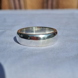 May include: A shiny, silver band ring. The ring has a smooth, rounded design and reflects the surrounding light. The ring is a simple, classic style, suitable for everyday wear. The ring is approximately 0.25 inches wide.