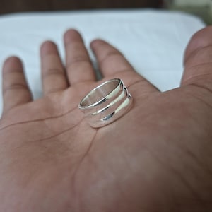 Puede incluir: Un anillo plateado brillante con un diseño de tres bandas. El anillo es liso y reflectante, captando la luz. La joya se muestra en una mano.