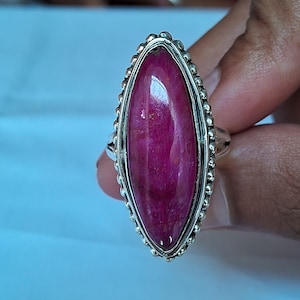 Puede incluir: Un anillo de plata con una gran piedra preciosa de color magenta en forma de marquesa alargada, engastada en un bisel. El engaste está enmarcado por un borde decorativo de pequeñas cuentas de plata, creando un borde texturizado.