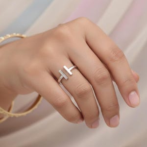 May include: A silver-colored ring with a modern design. The ring features two vertical bars that are connected by a thin band. The ring is worn on a finger, and the background is a soft, neutral color.