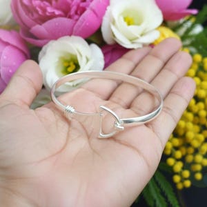 Könnte beinhalten: Ein silbernes Armband mit Hakenverschluss wird gezeigt. Das Armband ist rund und scheint aus poliertem Metall zu bestehen. Der Verschluss hat ein einzigartiges Design. Der Hintergrund umfasst bunte Blumen.