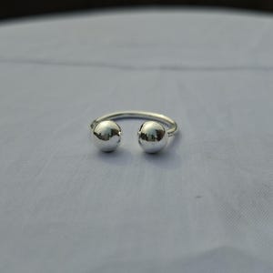 May include: A silver ring with two spherical accents. The ring has a simple, open design with a polished finish. The two spheres are positioned on either side of the open space, creating a minimalist and modern aesthetic.
