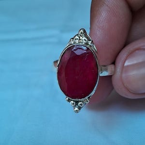 Puede incluir: Un anillo de plata con una gran piedra preciosa de color rojo intenso, tallada en forma ovalada y engastada en un marco de metal pulido. El engaste incluye detalles decorativos de cuentas de plata en los extremos superior e inferior.