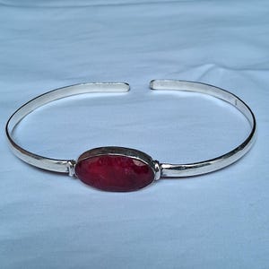 Puede incluir: Un brazalete de plata con una gran gema de rubí ovalada y facetada. El diseño abierto facilita su uso. El rubí es de un color rojo intenso, engastado en un bisel. Circunferencia aproximada de 17 cm.