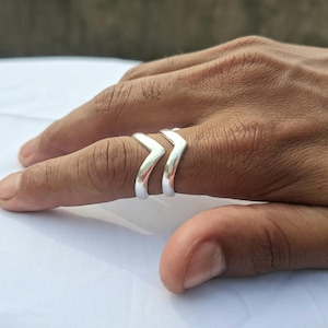 Puede incluir: Un par de anillos plateados con diseño de chevrón. Los anillos se lucen en un dedo, mostrando su superficie pulida y reflectante. El diseño en forma de V de los anillos añade un toque moderno a la joyería.