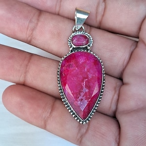 Könnte beinhalten: Silberanhänger mit einem großen, facettierten, tropfenförmigen rosa Edelstein. Oberhalb des Hauptsteins befindet sich ein kleinerer, ovaler rosa Stein. Der Anhänger hat eine dekorative, perlenbesetzte Bordüre und eine silberne Öse für eine Kette.