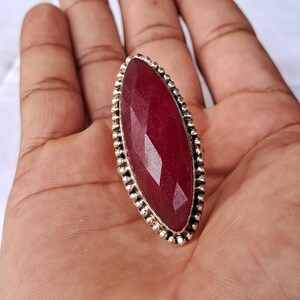Op de afbeelding: Een ring in marquisevorm met een grote, gefacetteerde dieprode edelsteen in een zilverkleurige metalen band. De zetting is omrand met een decoratieve kralenrand die de langwerpige steen omlijst, wat zorgt voor een opvallende, textuurrijke afwerking.