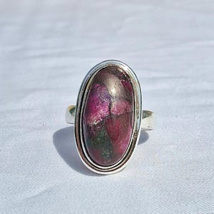 Puede incluir: Anillo de plata con una piedra preciosa ovalada en tonos rojo oscuro, morado y verde. La piedra está engastada en un bisel de plata de doble banda. La banda del anillo es de plata lisa. Una joya.