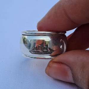May include: A wide silver band ring featuring a polished metallic finish and a detailed engraved illustration of a sailing ship on the front surface. The ring has a smooth, reflective texture with raised edges along the top and bottom borders.