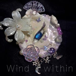 May include: A decorative, fantasy-themed art piece with a pearl-like flower, a blue eye, and a rose. The piece is adorned with butterflies, pearls, and charms. The text "Wind within" is at the bottom.