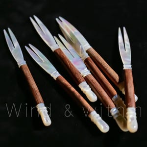 Puede incluir: Seis tenedores de ostras con mangos de madera, hojas de nácar y puntas blancas. Los tenedores están dispuestos sobre una superficie oscura. El texto "Wind & Within" es visible.