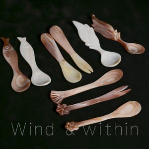 May include: A collection of decorative spoons and forks in various designs, including fish and wing shapes. The utensils are made of a pearlescent material with brown and white tones. The text "Wind & within" is at the bottom.