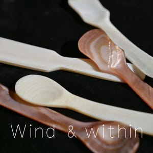May include: Close-up of several mother-of-pearl spoons in shades of white, cream, and pink. The spoons are arranged on a dark surface, with the text "Wind & within" in white at the bottom. The spoons have a smooth, polished finish.
