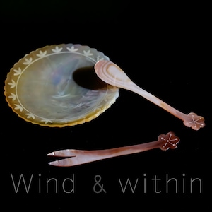Op de afbeelding: Een parelmoer serveerset met een geschulpte schaal, een lepel en een vork. De schaal heeft een bloemenpatroon langs de rand. De lepel en vork hebben bloemvormige uiteinden. De tekst "Wind & within" staat onderaan.