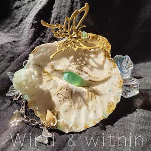 May include: An ornate, decorative seashell with a gold butterfly accent. The shell is white with gold and blue accents, a green gem, and a small chain with charms. The text "Wind & within" is at the bottom.