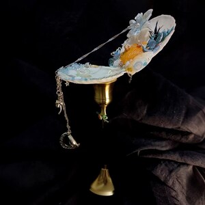 Puede incluir: Una campana de viento decorativa con una parte superior en forma de concha adornada con detalles florales azules y blancos, perlas y una campana dorada. Una cadena plateada con dijes, incluyendo una luna y un trébol de cuatro hojas, cuelga debajo.