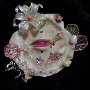 May include: A decorative seashell art piece featuring a central pink eye design with eyelashes. Adorned with pearls, stars, butterflies, and a large white flower, the piece has a whimsical, feminine aesthetic. The color palette includes pink, white, and iridescent accents.