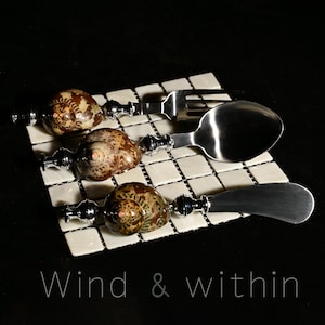 Puede incluir: Un juego de cuatro utensilios de acero inoxidable: un tenedor, una cuchara y un cuchillo de mantequilla, cada uno con un mango decorativo con un diseño de concha natural. Los utensilios descansan sobre una superficie de baldosas cuadradas. El texto "Wind & within" está en la parte inferior.