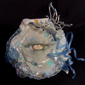 May include: A decorative, white, lip-shaped ornament with a silver butterfly accent. The ornament features blue and iridescent accents, including small butterflies, pearls, and a blue ribbon. The base is trimmed with lace.