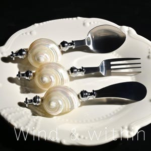 May include: A set of three pieces of cutlery: a spoon, fork, and butter knife. Each has a polished silver handle and a seashell-shaped grip. The cutlery rests on a white, scalloped-edge plate. The text "Wind & within" is at the bottom.