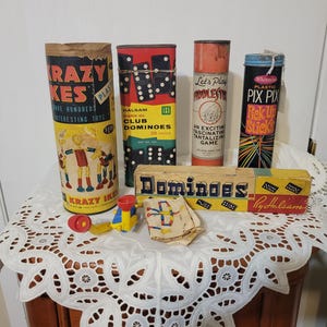 May include: A collection of vintage games, including dominoes, pick-up sticks, and a game called "Razy Ikes." The games are in their original packaging, with colorful graphics and text. The items are displayed on a white lace-covered table.