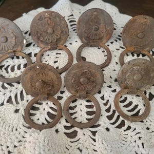 May include: A collection of seven antique bronze drawer pulls. Each pull features a circular, ornate face with a floral design and a hanging ring. The pulls are arranged on a white, open-weave surface.