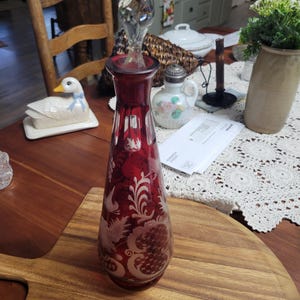 May include: A ruby red glass decanter with a clear glass stopper. The decanter features etched white floral and dragon designs. The decanter is approximately 12 inches tall.