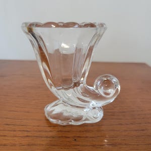 May include: Clear glass cornucopia-shaped vase with a scalloped rim and a curled, horn-like design. The vase sits on a decorative base and is displayed on a wooden surface. The vase is approximately 4 inches tall.