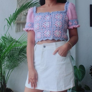 Puede incluir: Top de crochet en rosa pastel y azul con escote cuadrado y mangas abullonadas cortas. El top tiene una botonadura y un diseño floral de cuadrados de la abuela. Combinado con una falda de pana blanca.