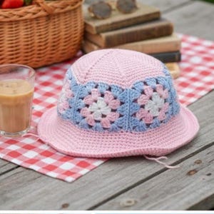 Puede incluir: Un sombrero de pescador rosa tejido a ganchillo con una banda con estampado floral azul y blanco. El sombrero está hecho de hilo suave y tiene un ala ancha. El sombrero está sobre una superficie a cuadros rojos y blancos.