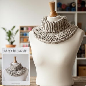 May include: A light gray roll neck cowl displayed on a mannequin. The cowl has a ribbed design at the neck and a textured pattern on the body. A card with the text "Knitt Fiber Studio" and "Roll Neck Cowl" is in the foreground.