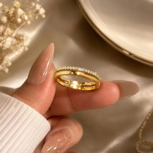 Peut inclure: Une bague dorée présentant un design à double anneau, dont l'un est lisse et poli, tandis que l'autre est incrusté d'une rangée de petites pierres claires et scintillantes qui captent la lumière, offrant une apparence raffinée.