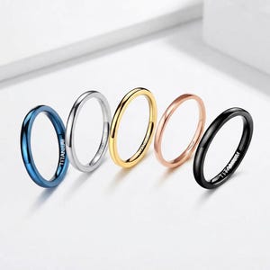 Pode incluir: Um conjunto de cinco anéis finos e lisos em azul, prata, ouro, ouro rosa e preto. Cada anel apresenta um acabamento polido e a palavra TITANIUM gravada na banda interna. Os anéis estão dispostos em fila, exibindo as suas cores metálicas.