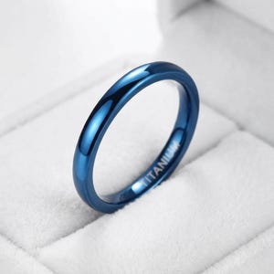 May include: A polished, deep blue ring with a smooth, rounded surface. The interior of the band features the word TITANIUM engraved in a clear, sans-serif font, indicating the material composition of this solid, circular jewelry piece.