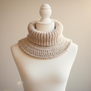 Op de afbeelding: Een beige gebreide colwarmer, gepresenteerd op een mannequin. De col heeft een geribbelde hals en een gestructureerd steekpatroon op het lichaam. De neutrale kleur en het eenvoudige ontwerp maken het een veelzijdig accessoire.