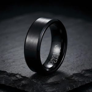 Könnte beinhalten: Ein schwarzer Ring mit gebürsteter Oberfläche und abgeschrägten Kanten. Auf der Innenseite des Rings befindet sich die Gravur TUNGSTEN CARBIDE. Der Ring hat ein massives, dunkles Erscheinungsbild mit glatter, metallischer Textur.