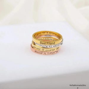 Peut inclure: Une pile de trois bagues fines en métal aux tons or, argent et or rose. Chaque bague présente un texte gravé, incluant les noms Ashley et Best Friend, ainsi que divers chiffres de coordonnées géographiques inscrits sur les anneaux.