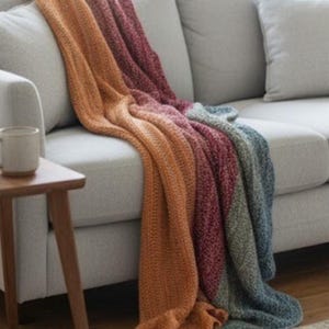 May include: A knitted throw blanket draped over a gray sofa. The blanket features a color gradient from orange to burgundy to gray and blue. A small wooden side table and a mug are visible in the image.