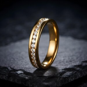 Peut inclure: Une bague anneau dorée dotée d'un sertissage rail avec une rangée continue de petites pierres claires taillées en rond incrustées au centre. La surface métallique présente une finition polie et réfléchissante, soulignant la forme circulaire.