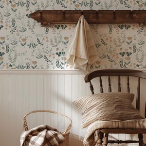 May include: A cozy interior scene featuring a wooden coat rack with a cream-colored towel, a wicker basket with a plaid blanket, and a wooden chair with a knitted pillow and throw. The wallpaper has a floral pattern in muted tones.