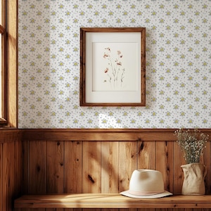 May include: A framed artwork featuring a botanical illustration hangs on a wall covered in a floral wallpaper pattern. A straw hat with a pink band sits on a wooden shelf. A vase of dried flowers is also on the shelf.