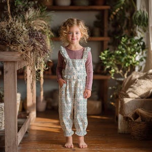 May include: A young child wearing a long-sleeved mauve top and a white jumpsuit with a blue floral pattern. The jumpsuit has ruffled straps and a gathered waist. The child is standing in a room with wooden shelves and plants.